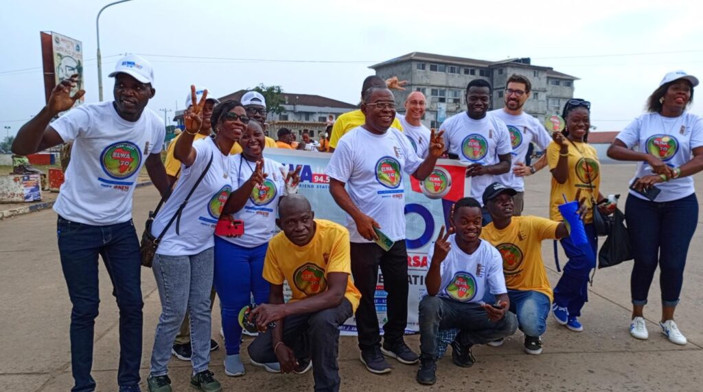 A group of people wearing white and yellow t-shirts with a colourful logo, posing for a photo outdoors in front of a banner