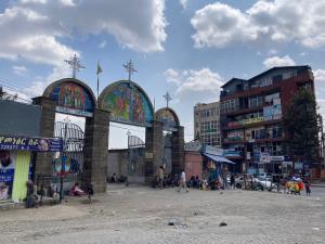 A street scene with an arched entrance featuring three arches topped with crosses, colorful religious artwork, people sitting and standing around, and buildings in the background under a partly cloudy sky.