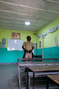 A person stands in a classroom with green walls, a whiteboard, and desks arranged in rows.
