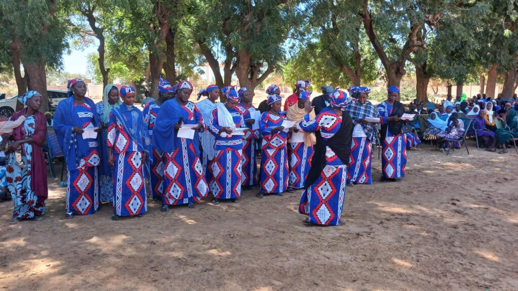 A group of people dressed in colorful, traditional clothing with blue, red, and white diamond patterns stand in a line outdoors, with trees providing shade in the background.