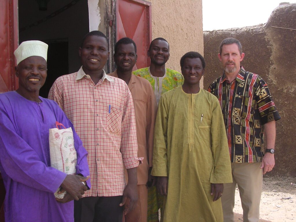 Six people stand in front of a building with an open door, wearing various traditional and modern clothing, including patterned shirts and long garments.