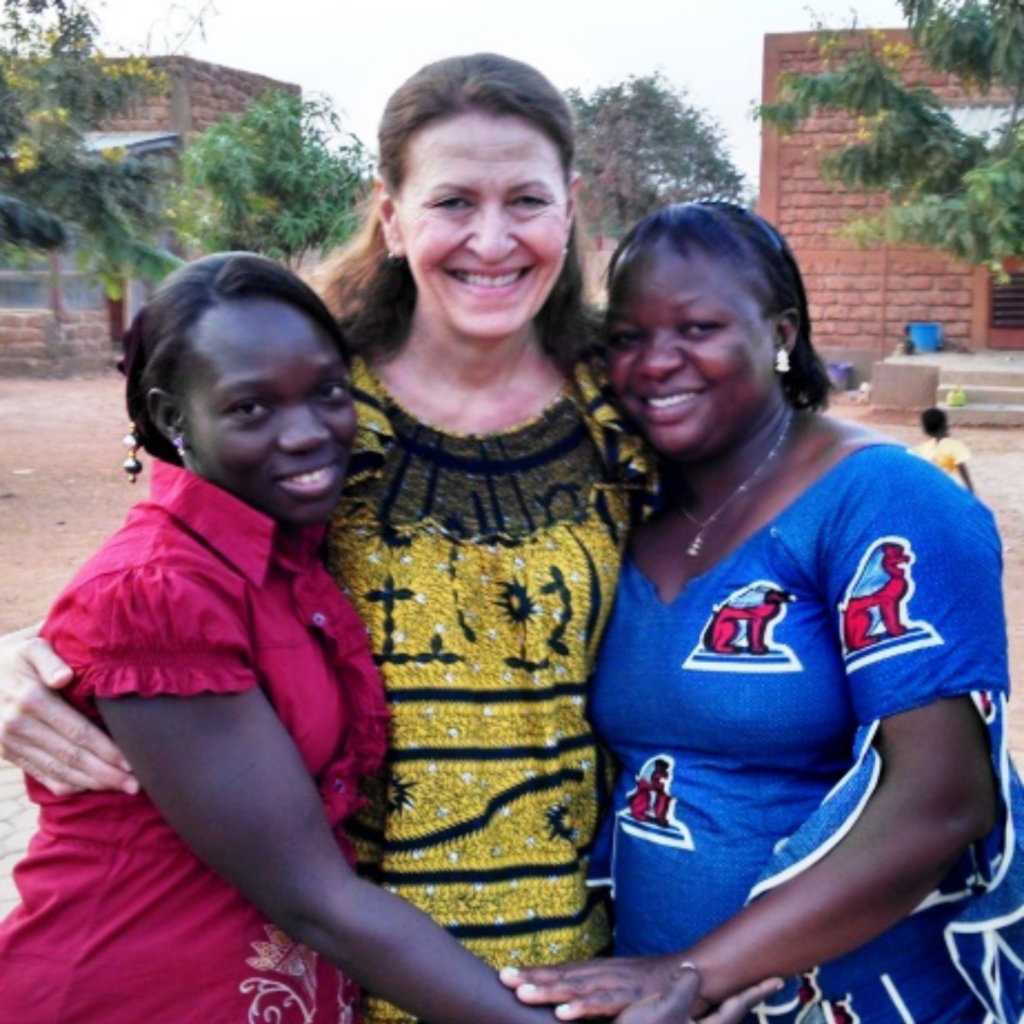 Three people stand close together outdoors; the person in the middle wears a yellow and black patterned dress, the person on the left wears a red top, and the person on the right wears a blue dress with red and white patterns.