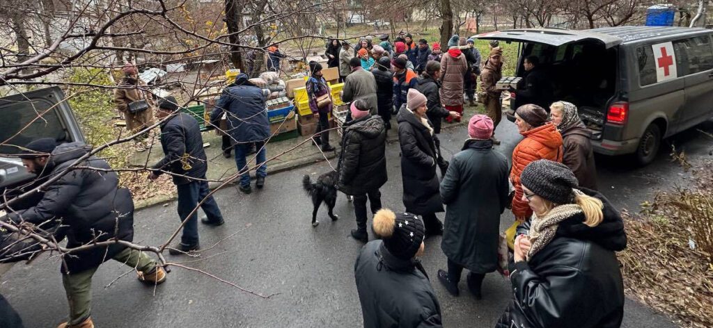 A group of people in winter clothing gathered around a van with a red cross symbol, distributing supplies outdoors.