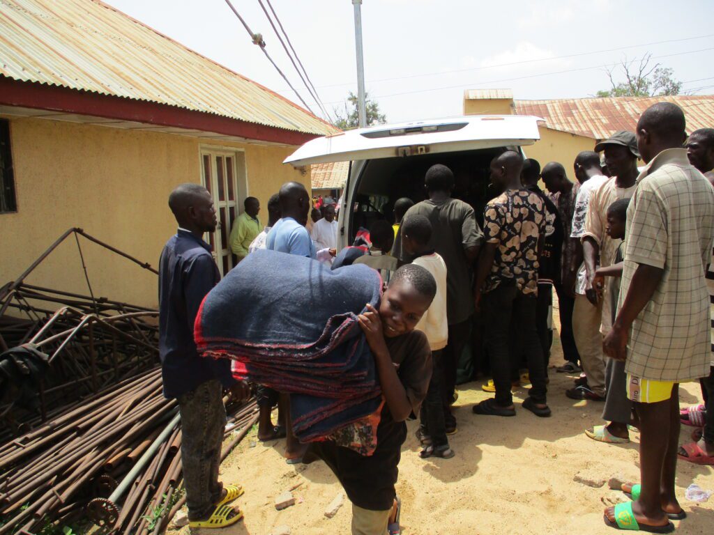 A group of people gathered around a vehicle, with one person in the foreground carrying a stack of blankets on their shoulder, outdoors near buildings and metal structures.
