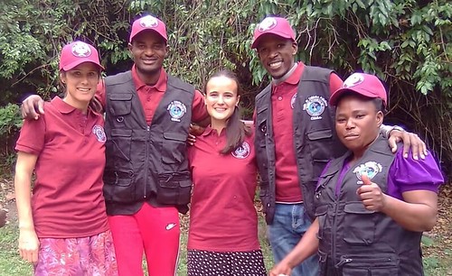 Five people standing together outdoors, all wearing maroon shirts and red caps with a logo, with three also wearing black vests with patches, against a background of green foliage and trees.