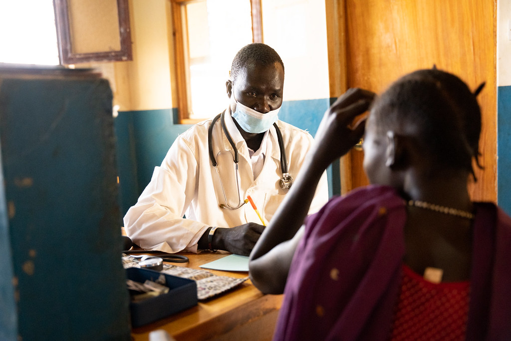 A man wearing a face mask with a stethoscope around his neck. In the foreground is a woman wearing bright clothing.