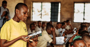 A classroom with students seated at desks and a person holding an open book in the foreground.